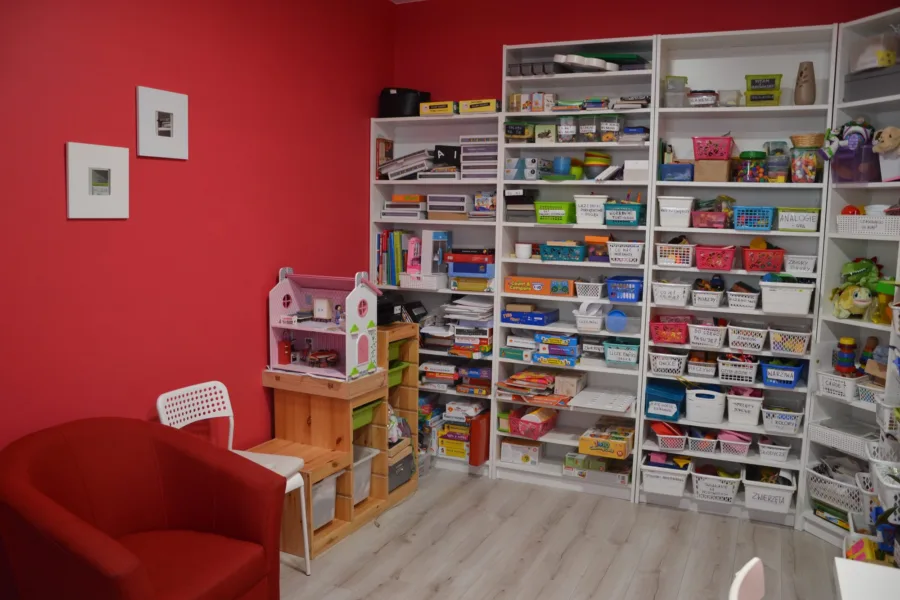 Sala terapeutyczna gabinetu: czerwona ściana, regały z pomocami logopedycznymi i fotel