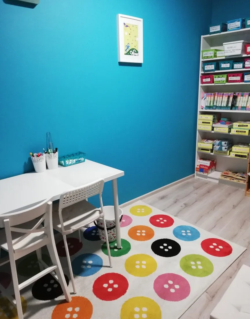 Sala terapeutyczna z turkusową ścianą, białym stolikiem i dywanem w kolorowe guziki