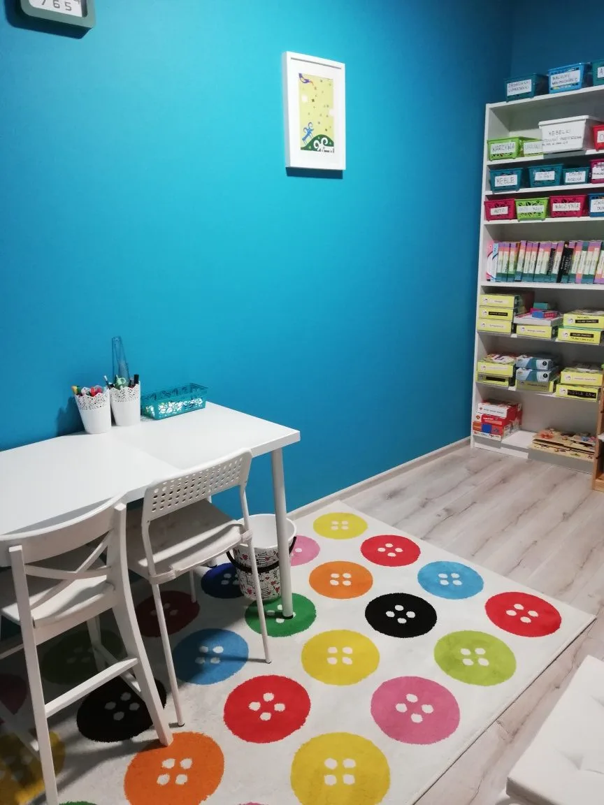 Sala terapeutyczna z turkusową ścianą, białym stolikiem i dywanem w kolorowe guziki